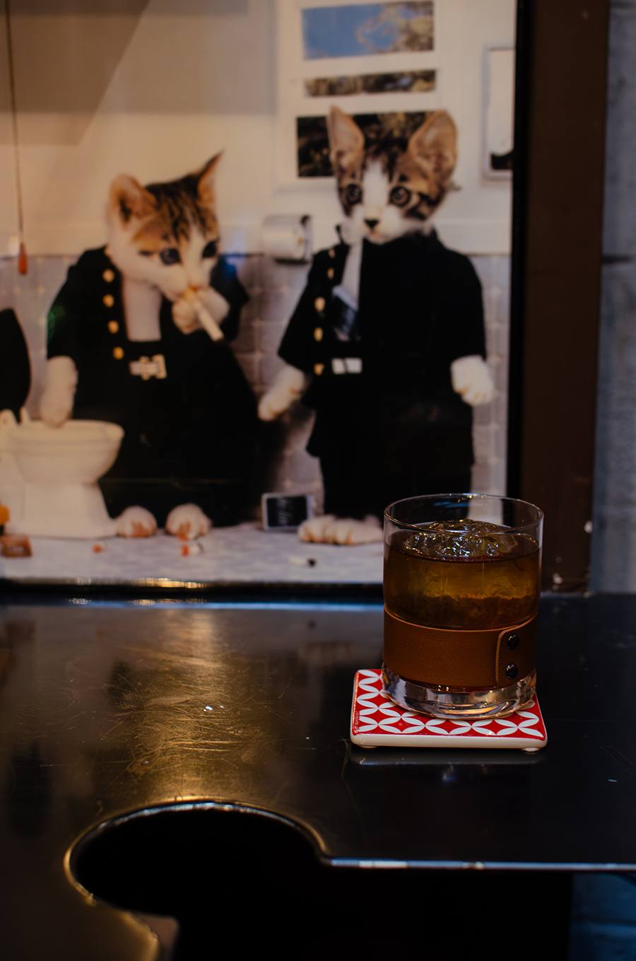 A glass of amber-colored drink with ice sits on a red and white patterned coaster on a dark reflective surface. In the background, there is a framed picture of two cats dressed in black outfits, one holding a cigarette near a toilet.