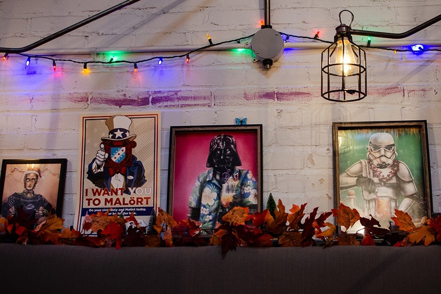 Four framed posters are displayed on a white brick wall. From left to right: a portrait of a person with glasses and a patterned shirt, a parody of the Uncle Sam "I Want You" poster with the text "I WANT YOU TO MALÖRT," a figure resembling Darth Vader wearing a colorful Hawaiian shirt, and a stormtrooper with a flower lei. Above the posters, colorful string lights and a cage-style hanging light fixture are visible. Autumn leaves decorate the surface below the posters.