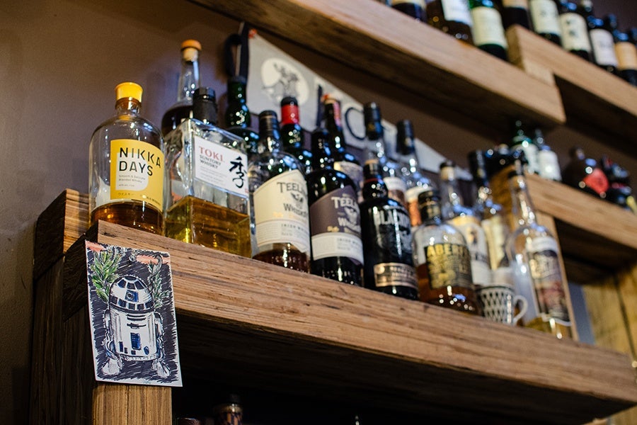 Wooden shelves holding various bottles of whiskey and liquor, including brands like Nikka Days, Toki, and Teeling, with a small illustrated card of the Star Wars character R2-D2 attached to the side of the shelf.