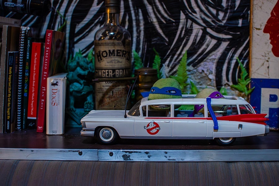 A white toy car modeled after the Ghostbusters Ecto-1, featuring the Ghostbusters logo on the door. Inside the car are two plush figures resembling Teenage Mutant Ninja Turtles, one with a blue mask and the other with a purple mask. The car is placed on a shelf with books and a vintage bottle in the background.