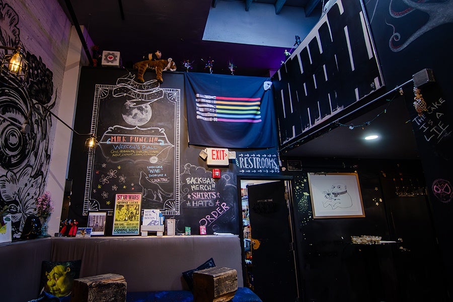 A cozy indoor space with dark walls decorated with various artworks and chalkboard signs. A large chalkboard features a moon illustration and text about "Milk Punch" and "Wrecking Ball." Below it, a smaller chalkboard advertises "Backbar Merch" including shirts and hats. A flag with colorful lightsaber designs hangs above an exit sign. The room has a mix of eclectic decorations, including a plush tiger on top of the chalkboard, a framed drawing of a frog on the right wall, and a mural of a large eye on the left wall. There are wooden stools and a cushioned bench with pillows, one featuring a Shrek design. String lights add a warm ambiance.