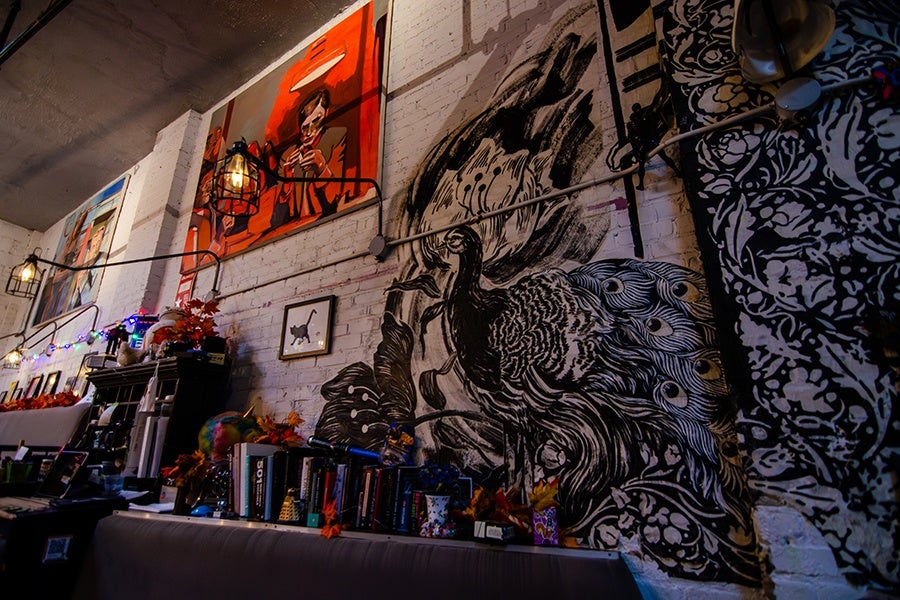 A wall with a large black and white mural of a peacock, surrounded by various framed artworks including a painting of a person on an orange background and a small framed picture of a cat. Below the mural, there are shelves filled with books, decorations, and autumn leaves, with string lights and industrial-style cage lamps hanging above.
