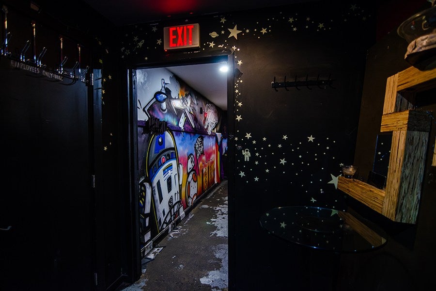A dimly lit hallway with black walls decorated with star and space-themed stickers, including a small astronaut figure. The open doorway reveals a colorful mural featuring Star Wars characters R2-D2 and BB-8. Above the doorway is a red illuminated "EXIT" sign. On the right side, there is a wooden-framed mirror and a small round glass table. The floor appears worn with patches of peeling paint.