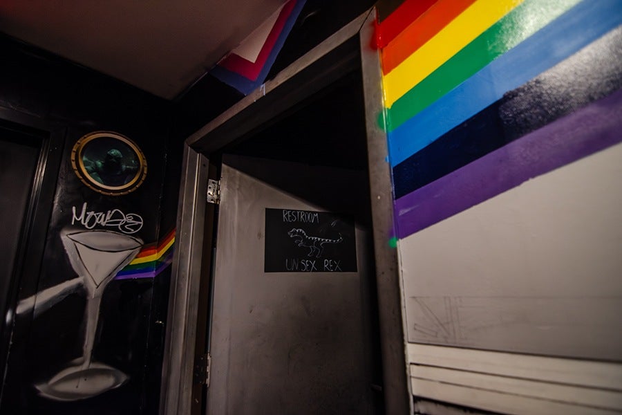 A partially open door with a sign that reads "RESTROOM UNISEX REX" and a drawing of a T-Rex. The surrounding walls are decorated with a rainbow stripe pattern and graffiti art, including a large martini glass.