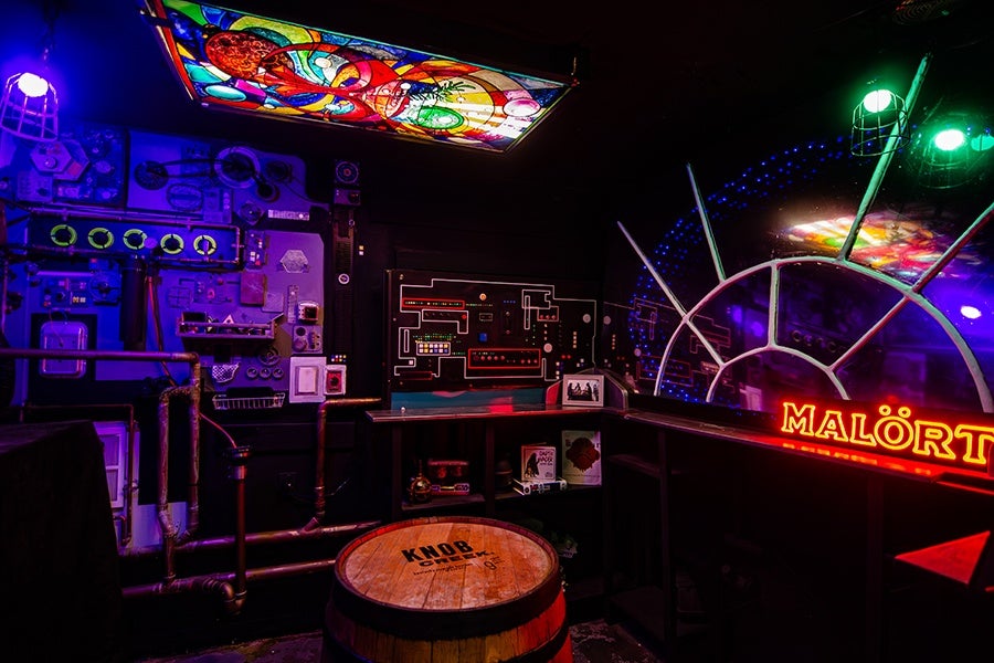 A dimly lit room with a stained glass ceiling light featuring colorful abstract designs. The walls are decorated with various electronic panels and circuits illuminated by blue and purple lighting. A wooden barrel with the label "KNOB" sits in the center. On the right side, a neon sign reading "MALÖRT" glows red, and there is a large circular window or mirror with metal framing and green lights above it. The overall atmosphere is industrial and futuristic.