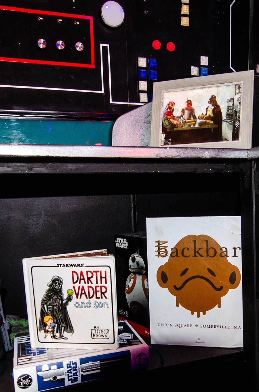 A framed picture shows Star Wars characters in a kitchen setting. Below it, there is a shelf with a Star Wars book titled "Darth Vader and Son" by Jeffrey Brown, a BB-8 toy box, and a white bag with an illustration of a sad-faced character and the text "ackbar" with the "B" crossed out, referencing Ackbar from Star Wars. The bag also mentions "Union Square, Somerville, MA." The background features a black panel with colorful buttons and dials.