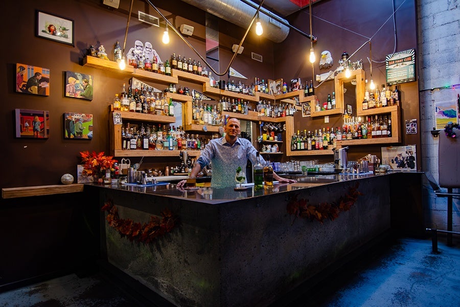 A bartender stands behind a modern, concrete bar counter decorated with autumn leaves. Behind him, a large wooden shelving unit displays a wide variety of liquor bottles. The walls are dark with colorful framed artwork and small decorations, including a pair of white stormtrooper helmets. Warm hanging light bulbs illuminate the space, creating a cozy atmosphere.