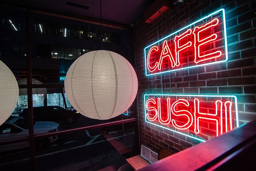 Red and blue neon signage on the brick wall of a restaurant reads Cafe Sushi, with a round white paper lantern hanging nearby.