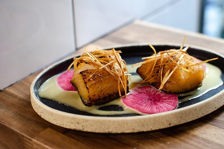 Two big pieces of fish, glossy and charred with a glaze, sit in an off-white pool of sauce next to pink slices of daikon.