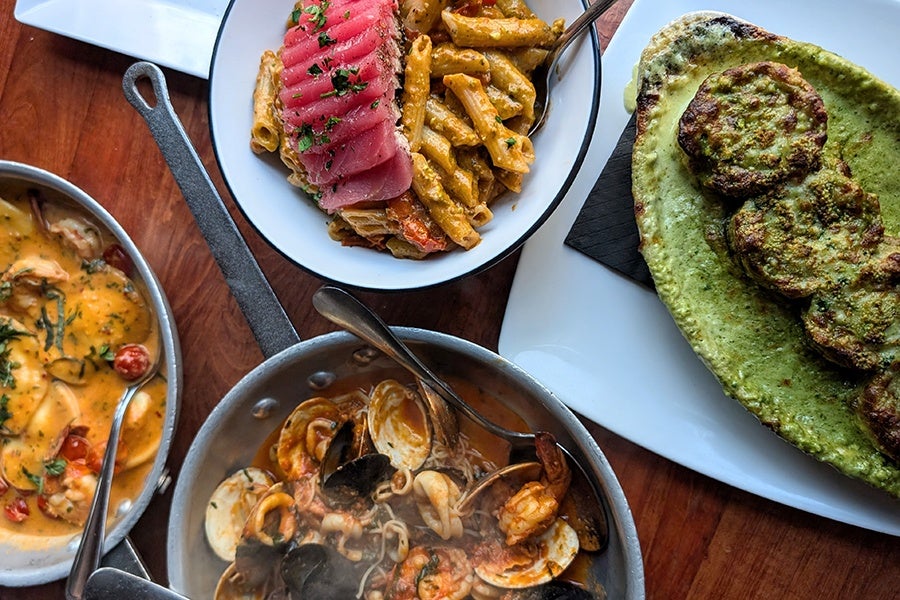 Overhead view of four pasta dishes on a wooden table, including penne topped with rare tuna, rolled up pasta sheets in a creamy green sauce, angel air with shellfish, and ravioli in a creamy orange sauce.