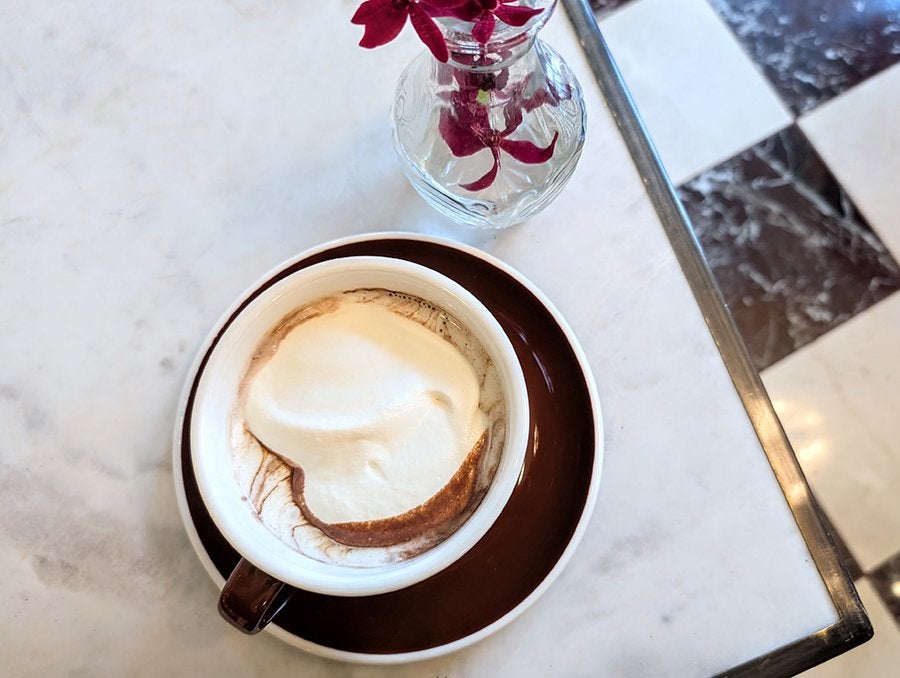 A cup of hot chocolate with whipped cream on top, served in a white cup with a dark brown saucer, placed on a white marble table. Next to the cup is a small glass vase containing a few dark red flowers. The table is positioned near a black and white checkered floor.