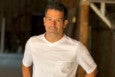 A man with short dark hair and light facial stubble is wearing a plain white short-sleeve shirt with a small pocket on the left side. He is standing indoors with a blurred background featuring wooden beams and a dimly lit space.