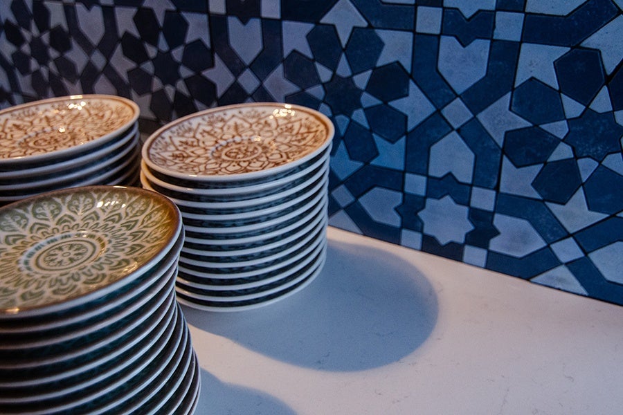 Stacks of small decorative plates with intricate patterns in green and brown, placed on a white countertop against a blue geometric patterned tiled wall.