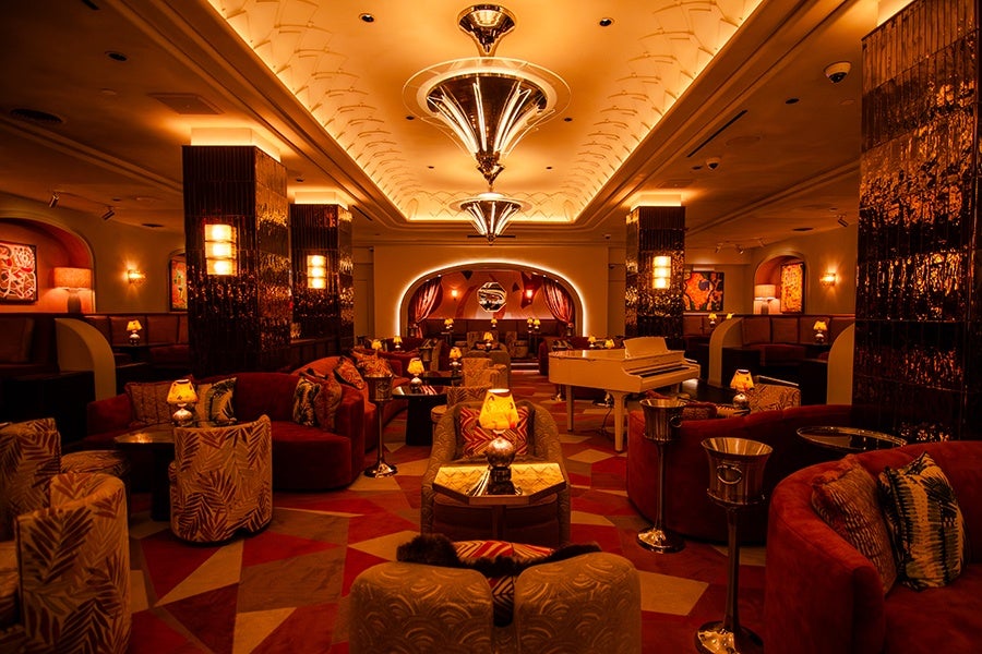 An upscale cocktail bar with a white grand piano and lots of red and gold tones and Art Deco vibes.