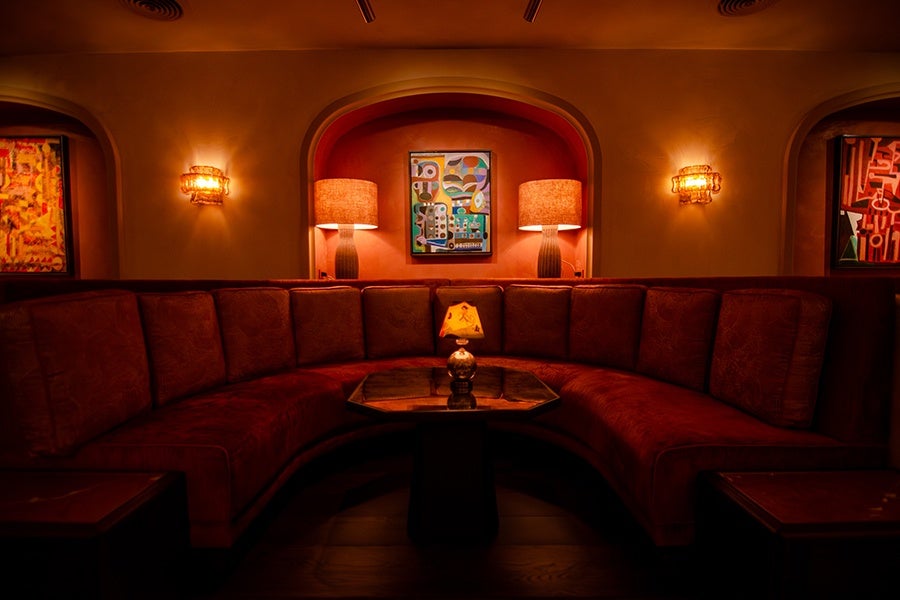 A curved red velvet couch in a dimly lit, upscale cocktail lounge, with colorful Cuban art hanging behind it.