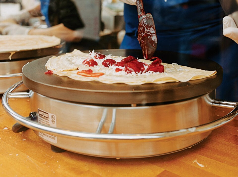 A crepe cooking on a round electric griddle with a person spreading a red sauce on it using a spatula. The crepe has some shredded white cheese and dollops of red sauce on top. The griddle is stainless steel with a black cooking surface and a protective metal ring around it. The person is wearing gloves and a blue apron.