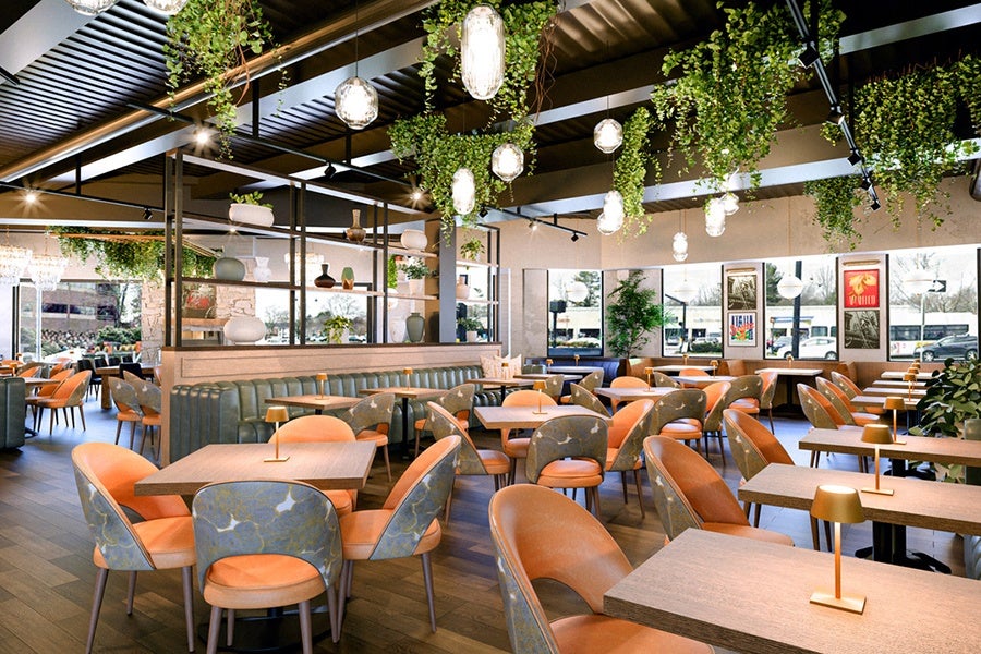 Modern restaurant interior with wooden tables and orange cushioned chairs featuring floral-patterned backs. Green hanging plants and pendant lights decorate the ceiling. The space includes a mix of booth seating with green upholstery and open chairs. Large windows along one wall allow natural light, and framed artwork is displayed on the walls. The floor is wooden, and small gold table lamps are placed on each table.