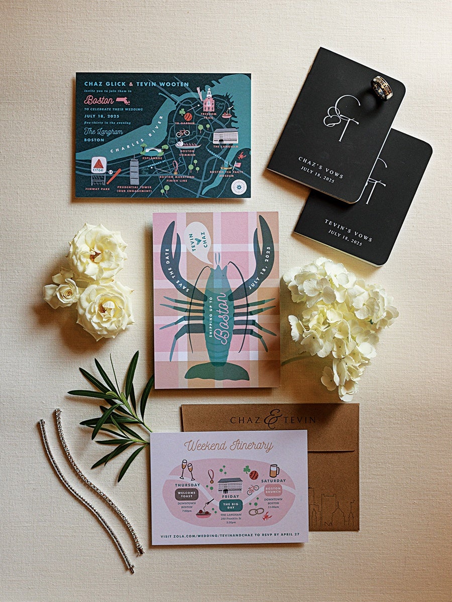 Wedding stationery set featuring a map-themed invitation with Boston landmarks, a pink and blue lobster-themed save-the-date card, two black vow booklets labeled "Chaz's Vows" and "Tevin's Vows" dated July 18, 2025, a brown envelope with "Chaz & Tevin" printed on it, and a weekend itinerary card with illustrated events. The set is accented with white flowers, green leaves, two silver bracelets, and a wedding ring placed on one of the vow booklets.