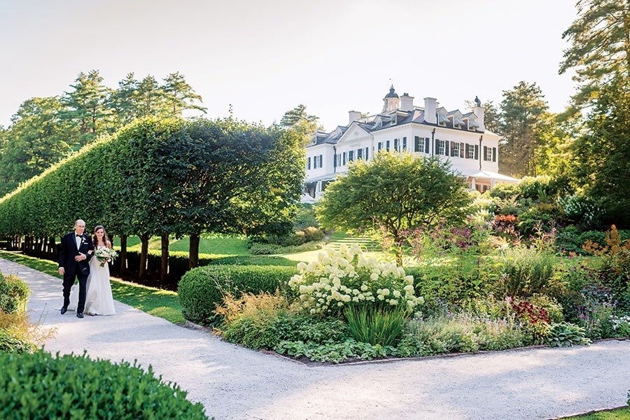 A bride in a white wedding dress holding a bouquet walks arm-in-arm with a man in a black tuxedo along a gravel path. They are surrounded by neatly trimmed trees, lush green bushes, and a colorful flower garden. In the background, there is a large, elegant white mansion with black shutters, set against a backdrop of tall trees and clear sky.
