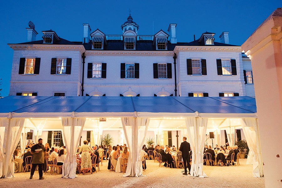A large white tent with sheer curtains is set up outside a grand white building with black shutters during dusk. Inside the tent, numerous people are seated at round tables, dressed formally, suggesting a social event or reception. Warm lights illuminate the tent, creating a cozy atmosphere against the deep blue evening sky.