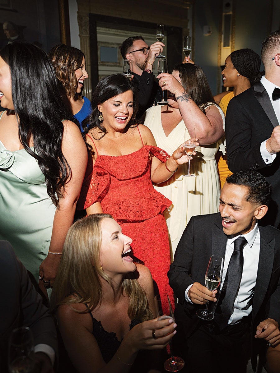 A lively group of well-dressed people at a celebration, holding champagne glasses and laughing. The central woman wears a red lace dress, surrounded by others in formal attire including a man in a black suit and tie, and women in dresses of various colors like light green, white, blue, and yellow. The atmosphere is joyful and festive.