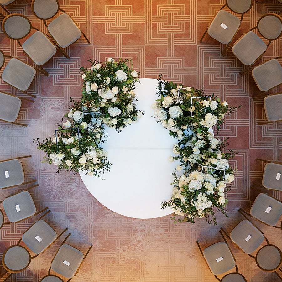 A round white table decorated with lush floral arrangements featuring white flowers and green foliage, surrounded by gray cushioned chairs arranged in a circular pattern on a patterned carpet. Each chair has a small white card placed on the seat.