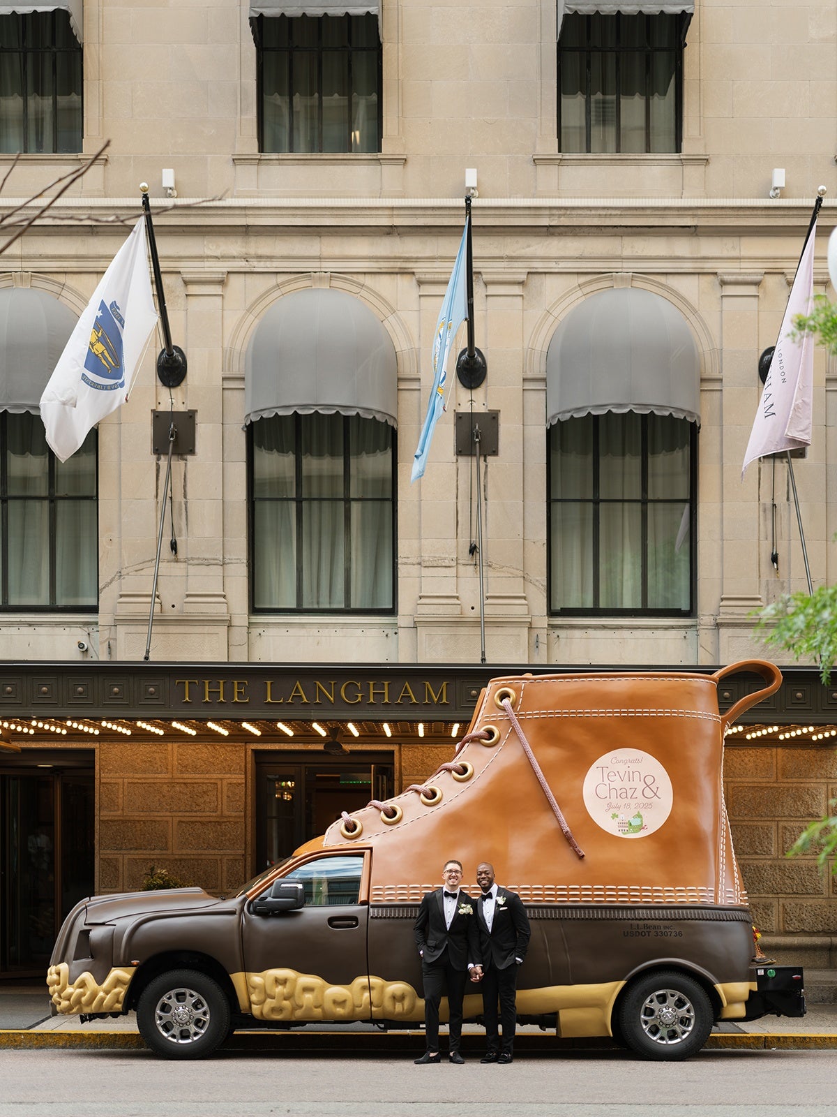 A brown and tan vehicle designed to look like a large boot is parked in front of a building with a sign that reads "THE LANGHAM." Two men in black tuxedos with bow ties and boutonnieres are standing in front of the vehicle, holding hands and smiling. The boot-shaped vehicle has a circular sign on the side that says "Congrats! Tevin & Chaz July 18, 2025." The building behind has beige stone walls, arched windows with gray awnings, and three flags mounted above the entrance.