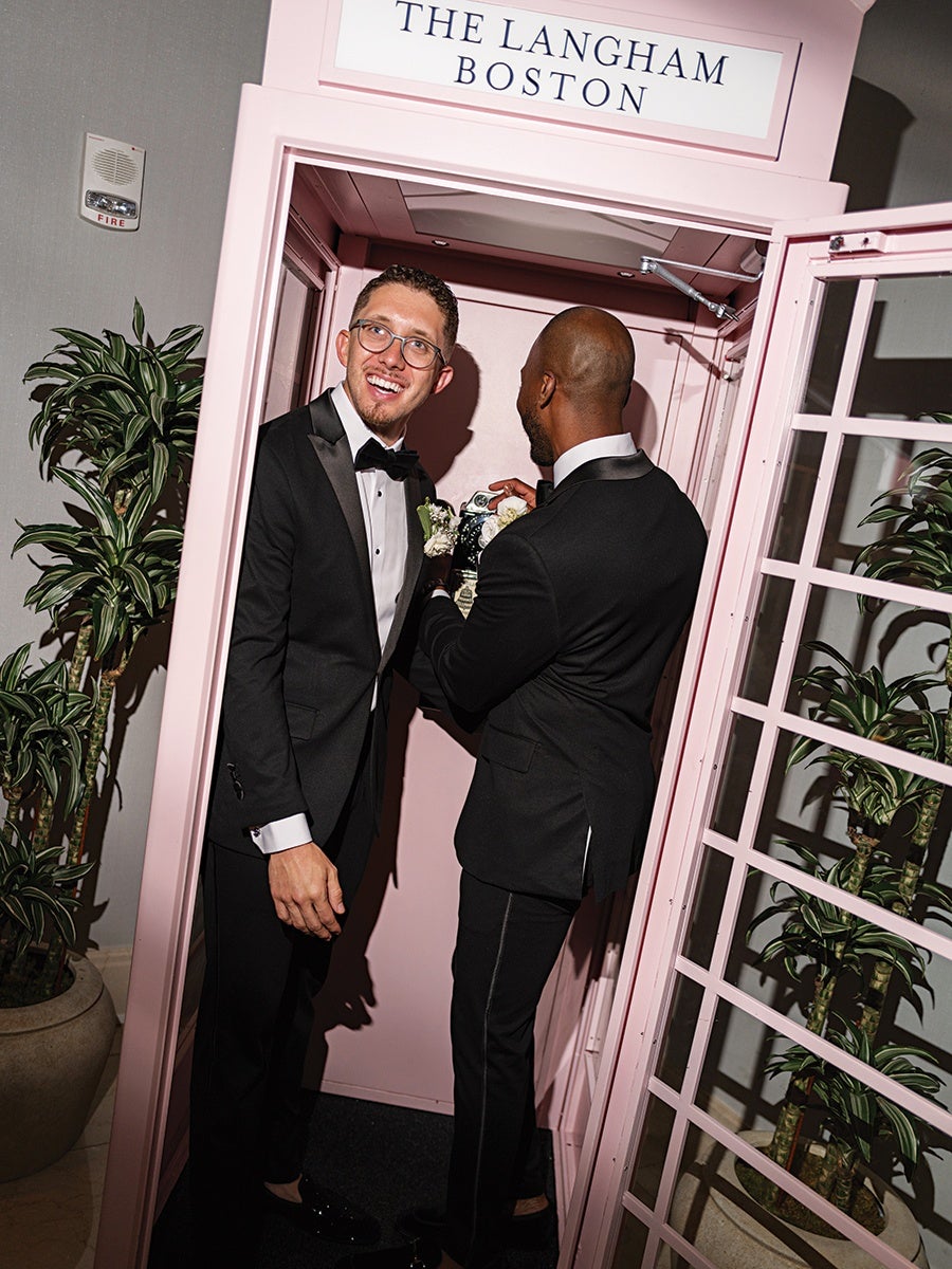 Two men in black tuxedos stand inside a pink phone booth labeled "The Langham Boston." One man, wearing glasses and smiling, faces the camera, while the other man, seen from the back, appears to be handling money. There are potted plants on either side of the phone booth.