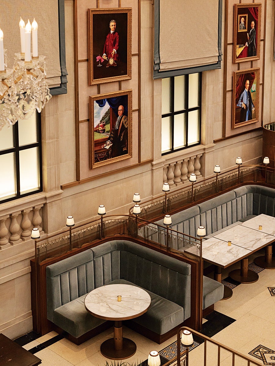 A sophisticated seating area with plush, gray velvet corner booths and marble-topped tables. The booths are separated by wooden dividers with small lamps on top. The walls are adorned with framed portraits of distinguished individuals, and large windows with beige Roman shades allow soft light to filter in. A crystal chandelier is partially visible on the left side, adding to the elegant ambiance.