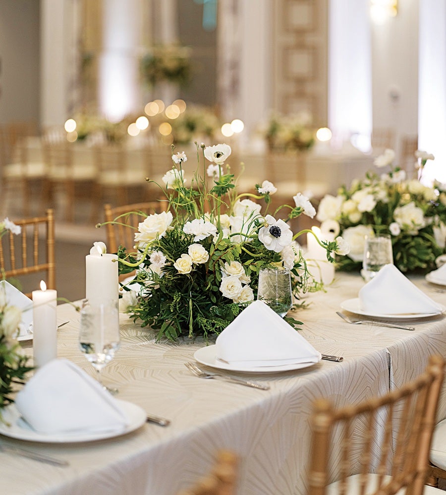 A formal dining table set with white plates, neatly folded white napkins, and clear water glasses. The table features elegant floral centerpieces with white flowers and green foliage. White candles in glass holders add a warm ambiance. The chairs are wooden with a light brown finish, complementing the sophisticated and serene setting.