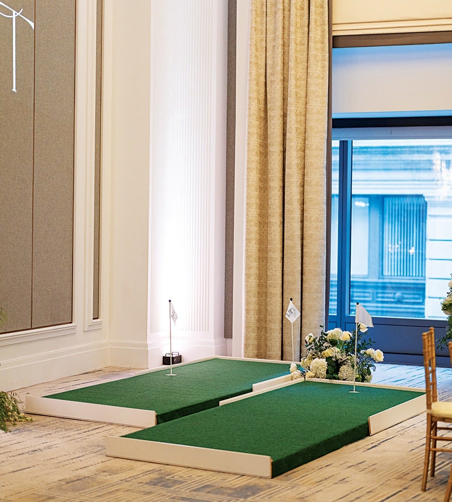 Two indoor mini golf putting greens with white borders and small flags, placed on a carpeted floor near a large window with beige curtains. There are floral arrangements near the greens and wooden chairs with yellow cushions nearby.