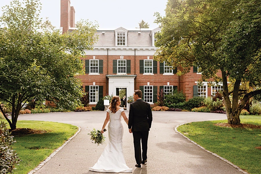 A bride and groom walk hand in hand down a paved driveway toward a large, elegant brick building with white trim and green shutters. The bride wears a white, off-the-shoulder wedding gown and holds a bouquet, while the groom is dressed in a black suit. The driveway is bordered by green grass and trees on both sides.