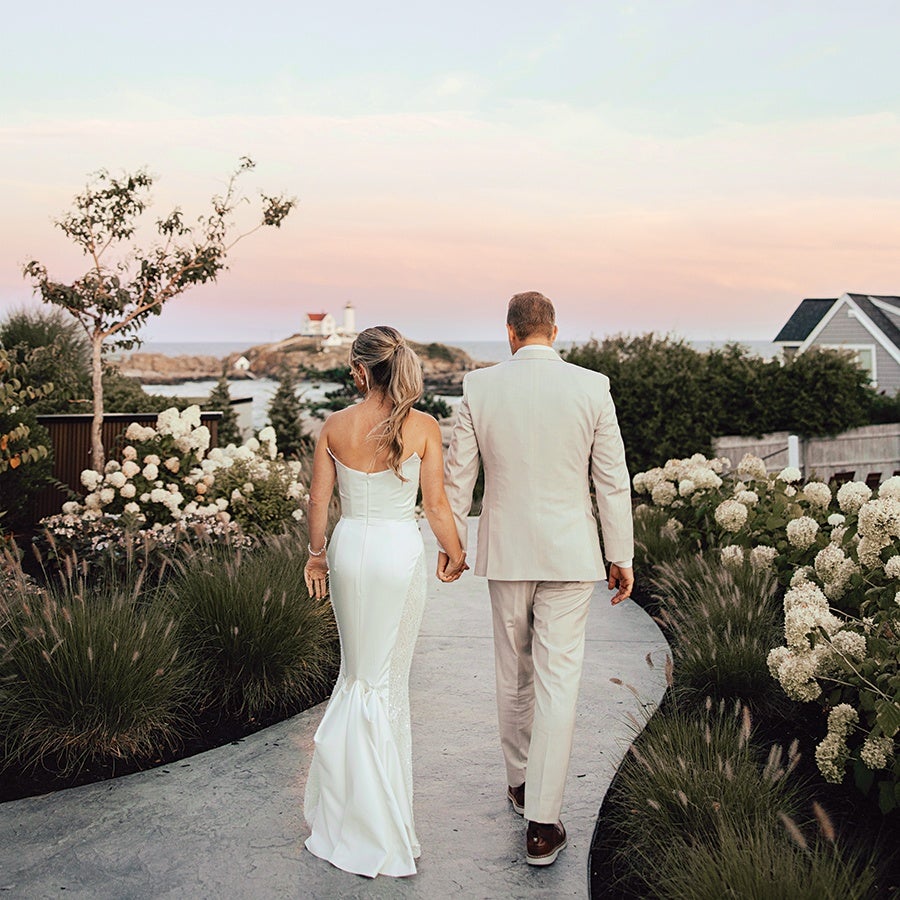 A couple dressed in wedding attire walking hand in hand on a curved garden path lined with white flowers and greenery, with a lighthouse and ocean visible in the background under a pastel sunset sky.
