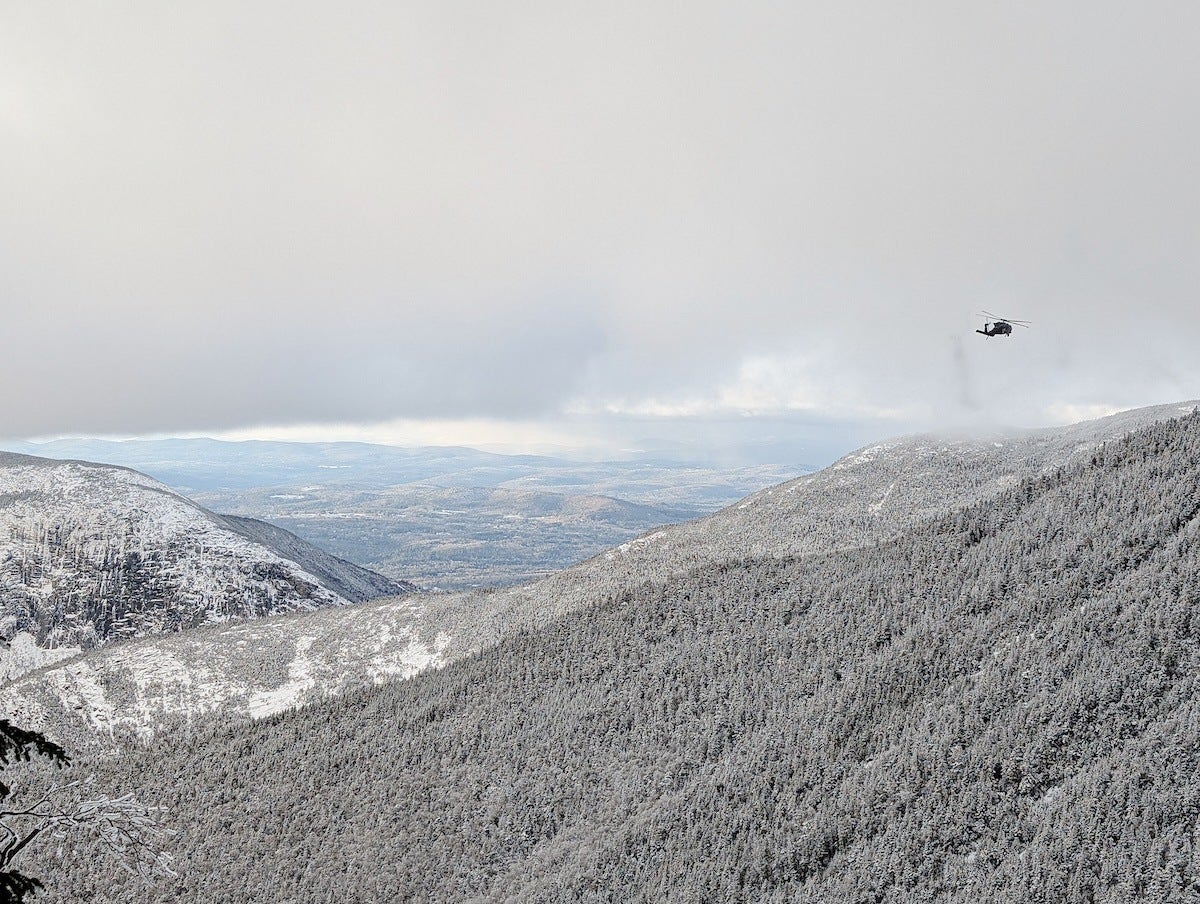 A snowy mountain landscape with dense forest covering the slopes. A helicopter is flying above the right side of the mountain under a cloudy sky. The distant horizon shows a mix of hills and flat land.
