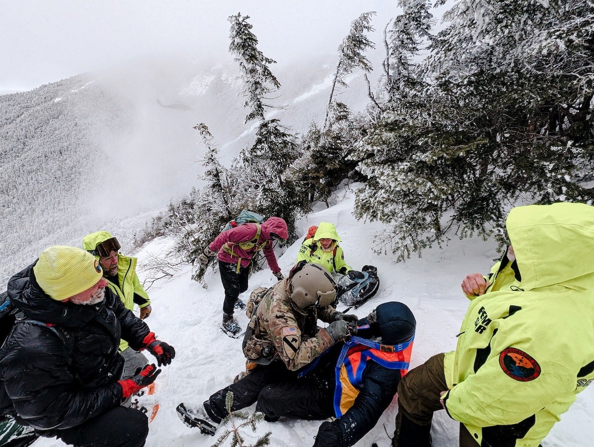 A group of people in winter gear are on a snowy mountain slope. One person in camouflage and a helmet is attending to another person lying on the snow wearing a blue and orange jacket. Others in bright yellow jackets and winter clothing are gathered around, some standing and some crouching, with snow-covered trees and a misty mountain in the background.