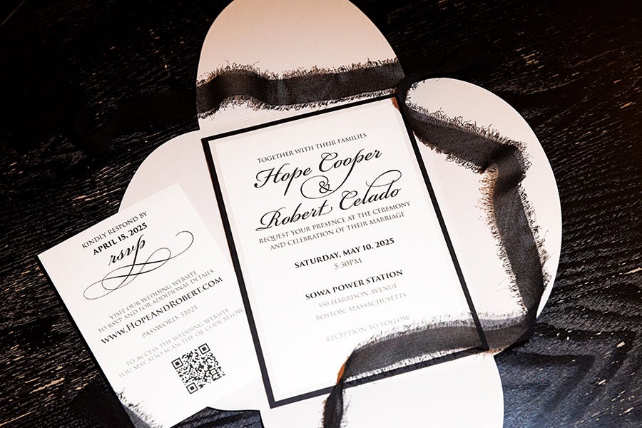 Wedding invitation set with a main card featuring the names Hope Cooper and Robert Celado, the date Saturday, May 10, 2025, and the venue Sowa Power Station in Boston, Massachusetts. The invitation is accompanied by an RSVP card with a website link and QR code, all tied together with a black sheer ribbon on a dark wooden surface.