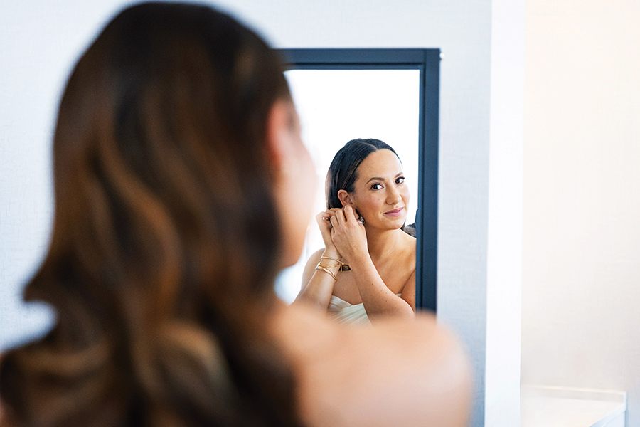 A woman with long, wavy brown hair is seen from behind as she looks into a mirror, adjusting an earring. She is wearing a strapless white top and gold bracelets on her wrist. The background is minimal and light-colored.