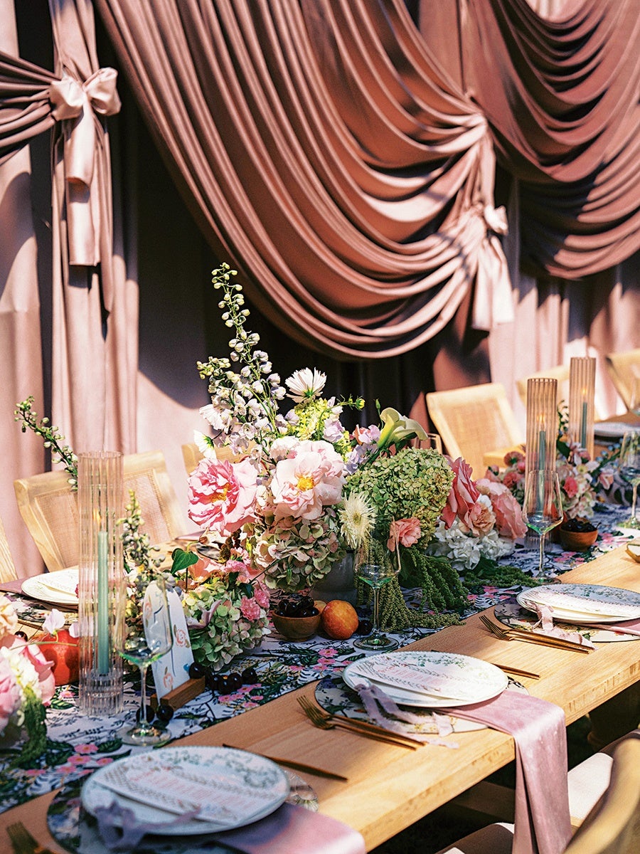 A wooden dining table set for a formal event with floral-patterned plates, gold flatware, and light purple napkins. The table features a floral centerpiece with pink, white, and green flowers, along with small bowls of fruit and olives. Tall, ribbed glass candle holders and wine glasses are also arranged on the table. In the background, there are elegant, dusty pink draped curtains tied with bows, and light wooden chairs with woven backs.