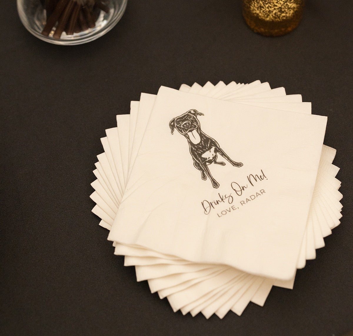 A stack of white napkins with a black illustration of a happy dog with its tongue out. Below the dog, the text reads "Drinks On Me! LOVE, RADAR" in a mix of cursive and uppercase letters. The napkins are placed on a dark surface.