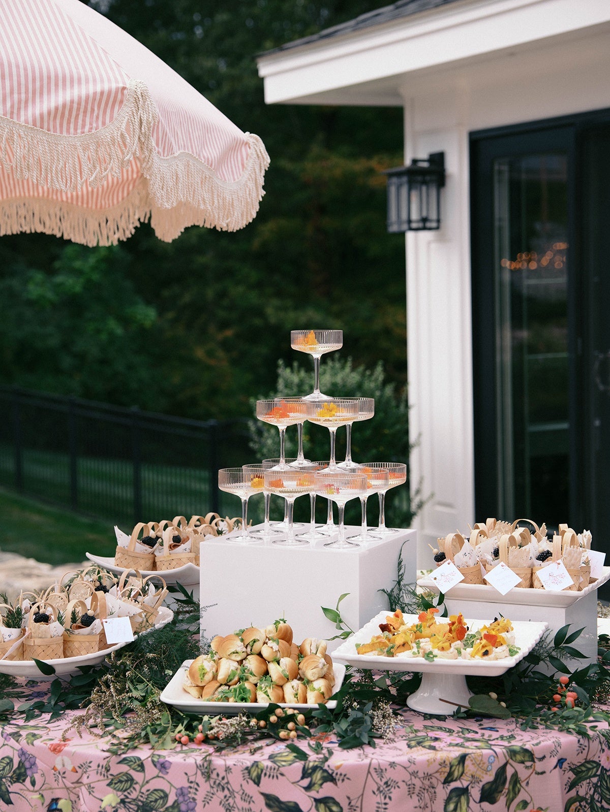 A table covered with a pink floral tablecloth displays a variety of appetizers and drinks. There is a pyramid of clear coupe glasses filled with a light-colored beverage and garnished with orange peel. Surrounding the glasses are plates of small sandwiches and bite-sized appetizers, some served in small woven baskets with white napkins and blackberries. Greenery and small flowers are used as decorative accents on the table. A pink and white striped umbrella with fringe hangs above the setup. The background shows a white building with a black door and outdoor lighting.