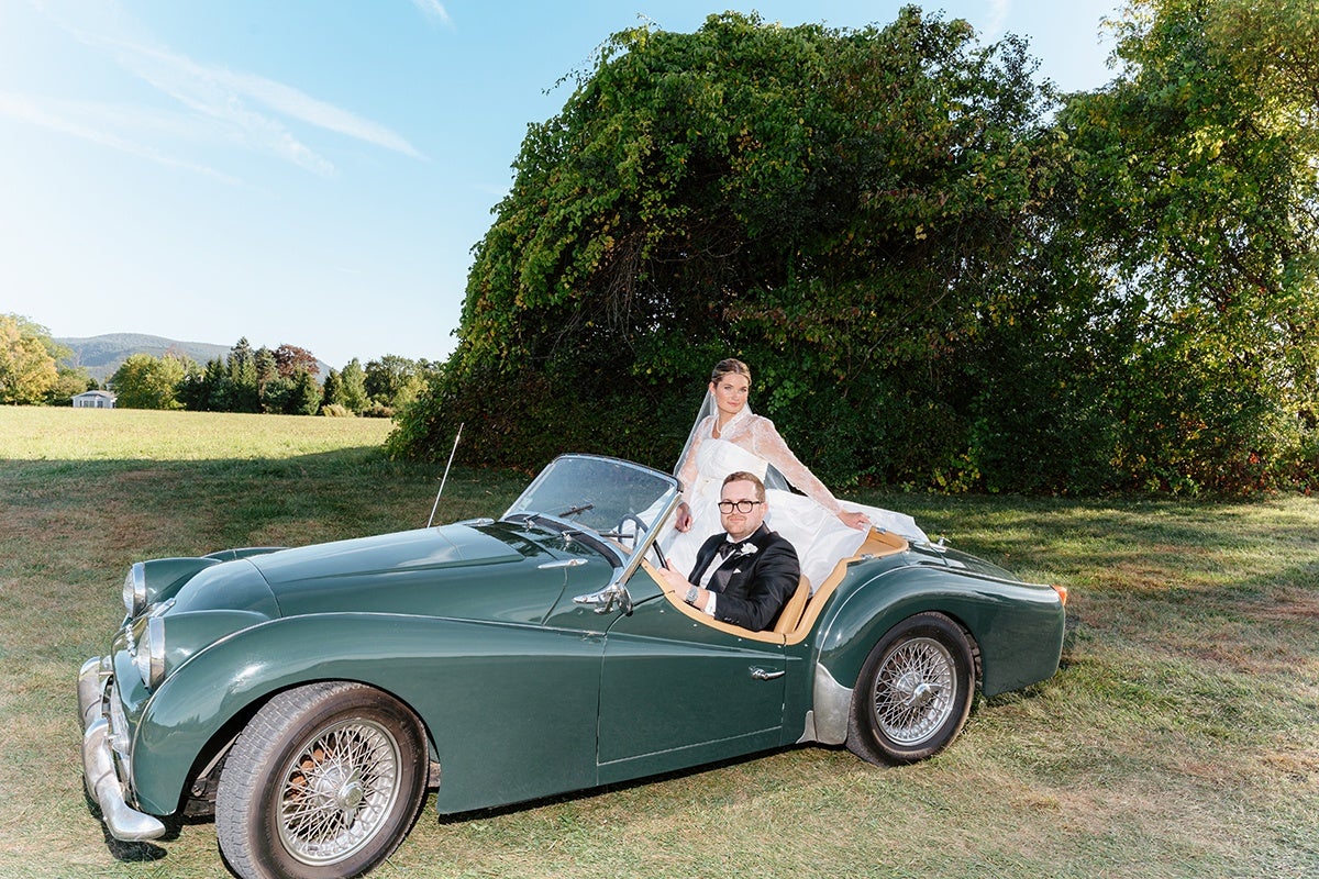 A bride in a white wedding dress and a groom in a black tuxedo are seated in a dark green vintage convertible car with tan interior, parked on grass with a large leafy bush and clear blue sky in the background.
