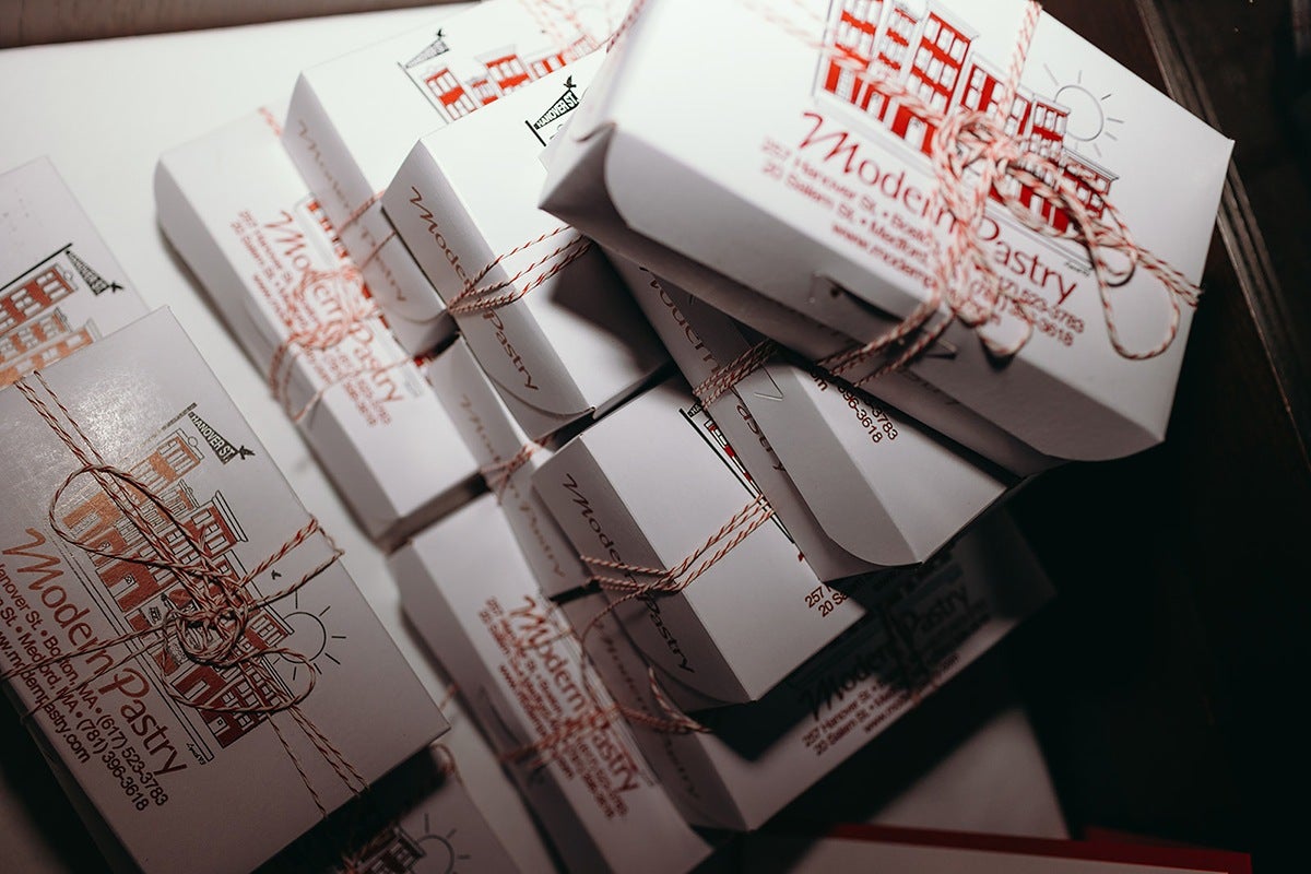 Stack of white pastry boxes tied with red and white twine, featuring a red illustration of buildings and the text "Modern Pastry" along with contact information. The boxes are arranged on a white surface with a dark background.