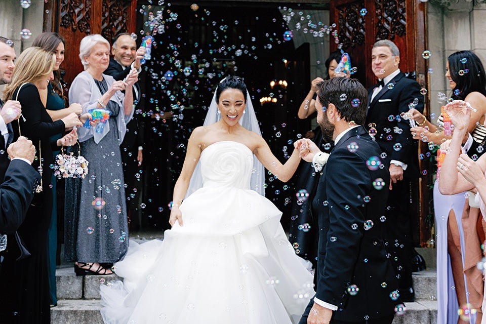 A bride and groom are standing outside a building, holding hands and smiling. The bride is wearing a strapless white wedding gown with a floral design on the bodice and a veil, while the groom is dressed in a black tuxedo with a white boutonniere. They are surrounded by guests who are blowing bubbles, creating a festive and joyful atmosphere. The guests are dressed in formal attire, and the background features ornate wooden doors.