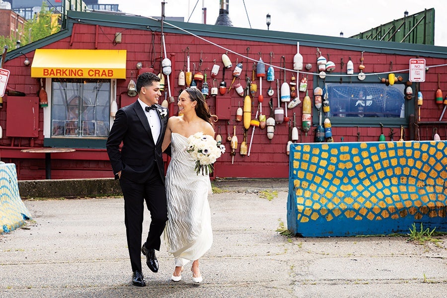 A bride and groom walking arm in arm outdoors in front of a red building with a yellow awning that reads "BARKING CRAB." The bride is wearing a strapless white wedding dress and holding a bouquet of white flowers, while the groom is dressed in a black tuxedo with a bow tie. The building's wall is decorated with colorful fishing buoys. Two blue and yellow painted barriers are visible on either side of the couple.