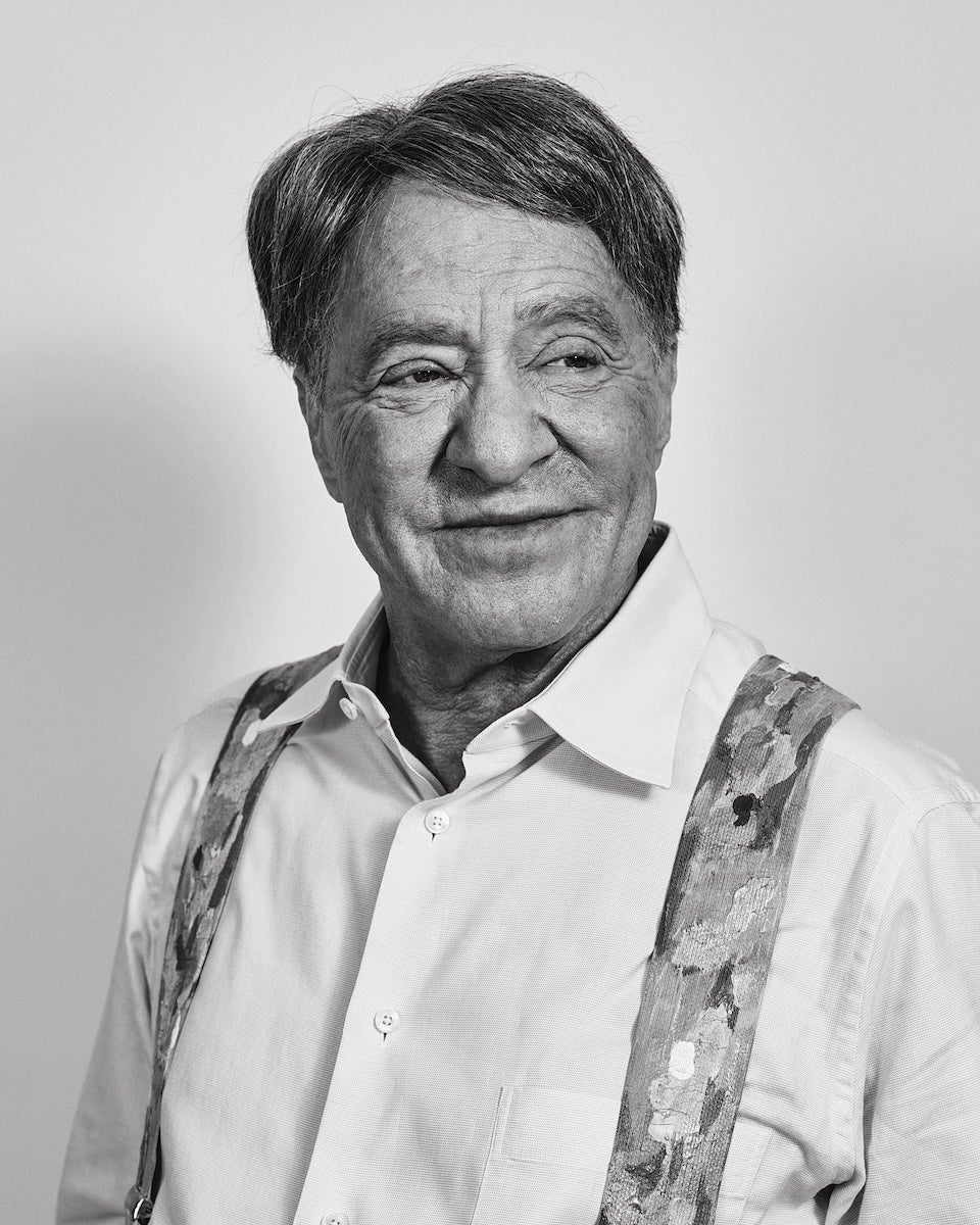 Older man with short, dark hair wearing a light-colored button-up shirt and patterned suspenders, looking slightly to the side with a gentle smile. The image is in black and white with a plain background.