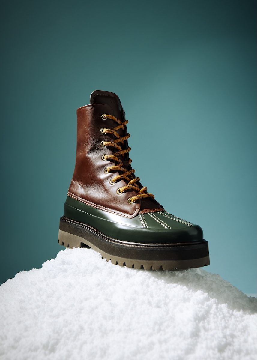 A high-top boot with a brown leather upper and dark green lower section, featuring yellow laces threaded through gold eyelets. The boot has white stitching details on the green part and a thick, rugged sole in dark brown and beige. It is displayed on a mound of white material against a teal background.