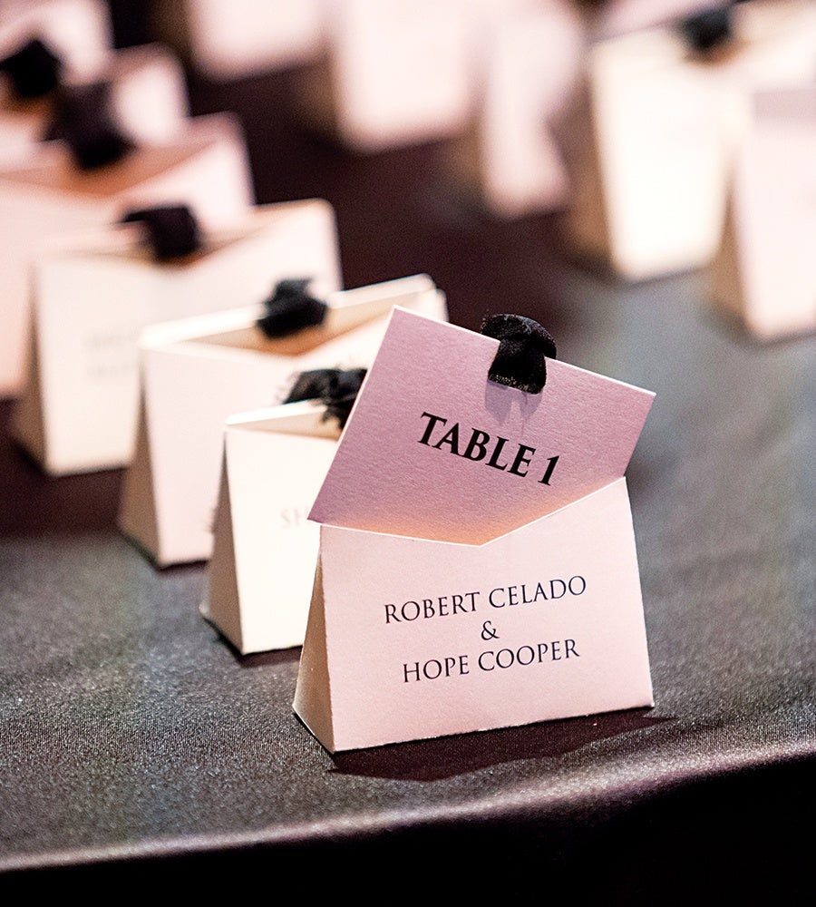 Folded white place card with black text reading "ROBERT CELADO & HOPE COOPER" and a smaller pink card inserted at the top with "TABLE 1" printed in black, set on a dark surface with similar place cards blurred in the background.