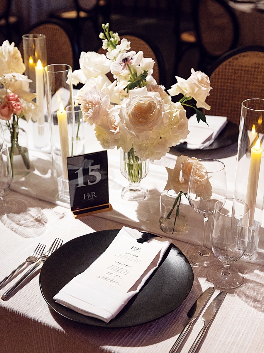 A formal table setting with a black plate holding a folded white napkin and a menu card. The table is decorated with white and pale pink flowers in glass vases, tall glass candle holders with lit candles, and a black table number card displaying "15." Silverware is neatly arranged on either side of the plate, and there are two clear glasses to the right. The tablecloth is white with a subtle texture.