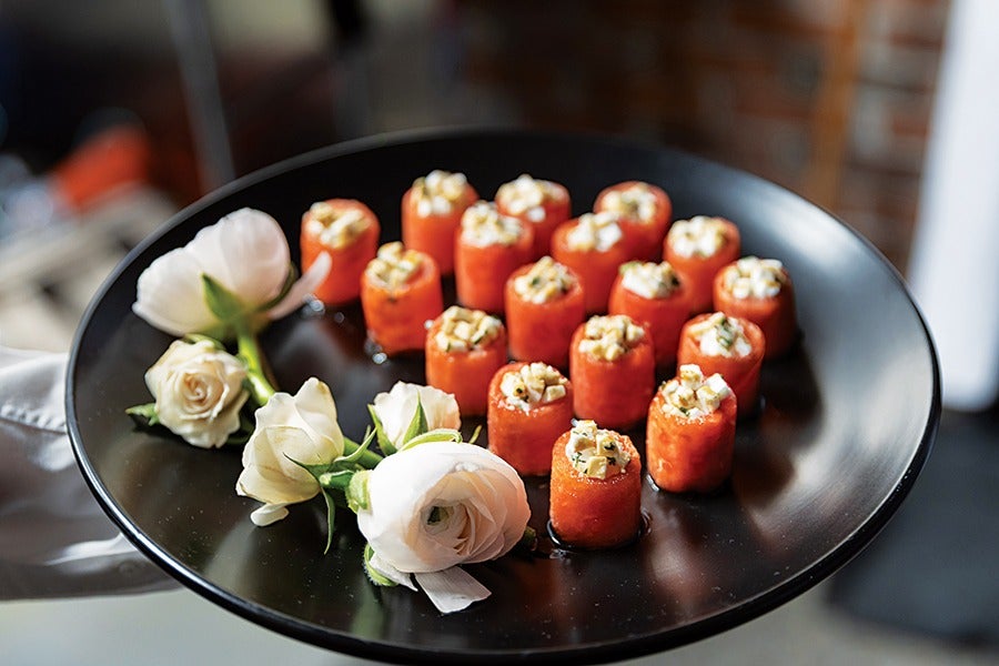 A black plate holds several small cylindrical appetizers with an orange outer layer and a creamy topping with herbs. The plate is also decorated with white and pale yellow roses.