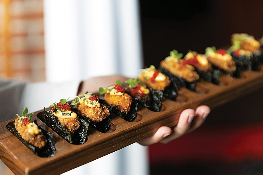 A wooden serving board holding a row of ten pieces of sushi, each wrapped in seaweed and topped with a crispy fried item, creamy sauce, red fish roe, and small green garnishes. A hand is holding the board.